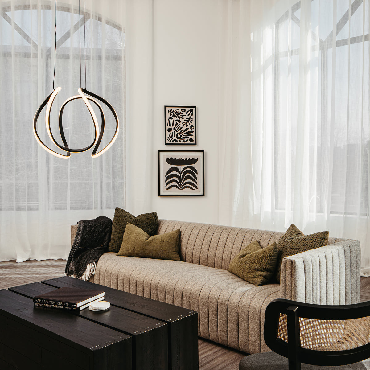 A modern living room with a striped beige sofa, green pillows, a black wooden coffee table, and abstract wall art. Sheer curtains cover large windows, and a sculptural pendant light hangs from the ceiling.