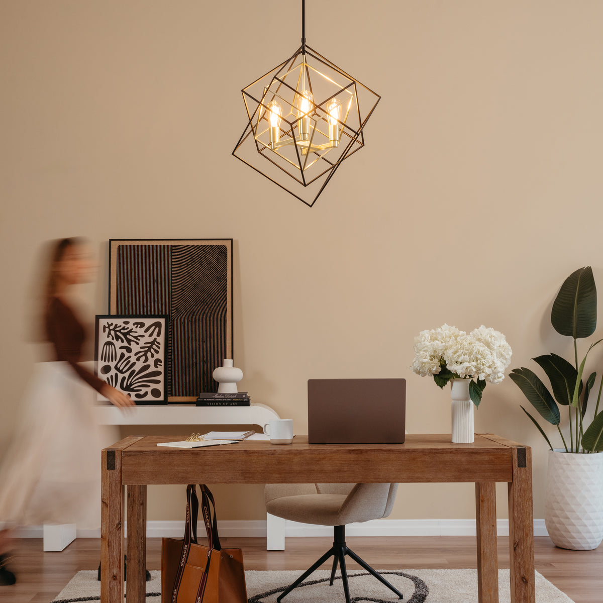 A modern home office with a wooden desk, laptop, coffee cup, vase of white flowers, and a tan chair. A blurred person walks by abstract art on the wall. A geometric pendant light hangs from the ceiling.