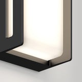 Close-up of a modern, geometric black wall sconce with soft white light glowing from within, mounted on a light gray wall. The fixture has clean, angular lines and a contemporary design.