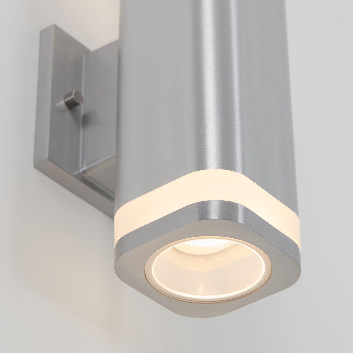 Close-up of a modern, wall-mounted cylindrical light fixture with a brushed metal finish and a glowing white strip near the bottom edge.