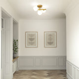 A hallway with light gray wainscoting, herringbone wood floor, two framed abstract art prints on the wall, a gold and white ceiling light, and a potted plant on a windowsill.