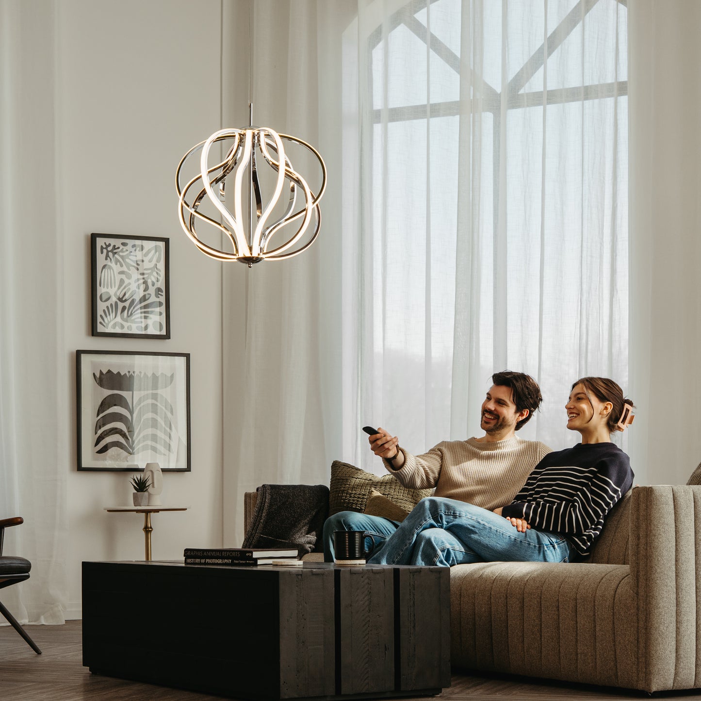A smiling couple relaxes on a beige sofa in a modern living room, watching TV. Light from the Artika Meridian LED pendant light streams through large windows, illuminating framed art on the wall.