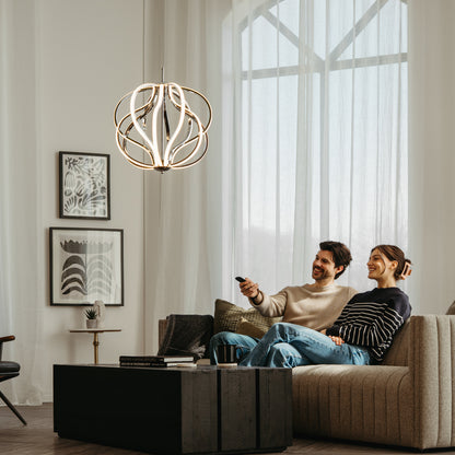 A smiling couple relaxes on a beige sofa in a modern living room, watching TV. Light from the Artika Meridian LED pendant light streams through large windows, illuminating framed art on the wall.