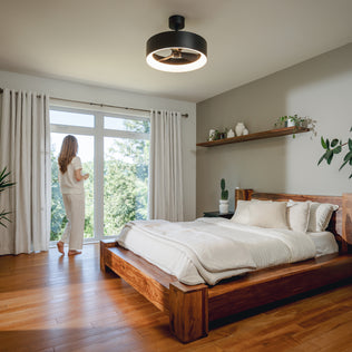 A woman stands in a bright room with plants, wooden floors, and an Artika Aiden 24 in. LED modern fandelier ceiling fan overhead.