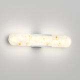 A modern wall sconce with a cylindrical, frosted, marble-like glass shade emits a soft, warm glow. The fixture is mounted horizontally with two sleek metal brackets on a light gray wall.