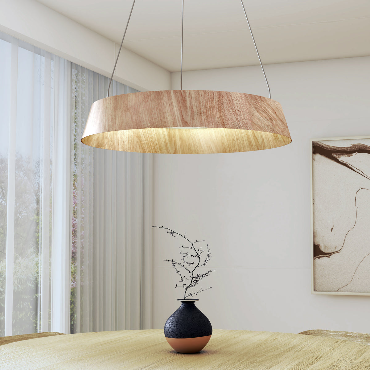 A modern dining room with a large wooden pendant light hanging above a round table. On the table is a dark vase with a slender branch, and behind is a window with sheer curtains and abstract wall art.