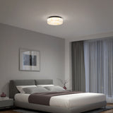 A modern bedroom with a gray upholstered bed, white bedding, a bedside table, a framed picture on the wall, large window with sheer curtains, and a round ceiling light fixture.