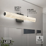 A modern wall light with a matte black finish and glass diffuser is mounted above a bathroom mirror. Text highlights its integrated LED, 5-year warranty, and feature of no bulbs required.