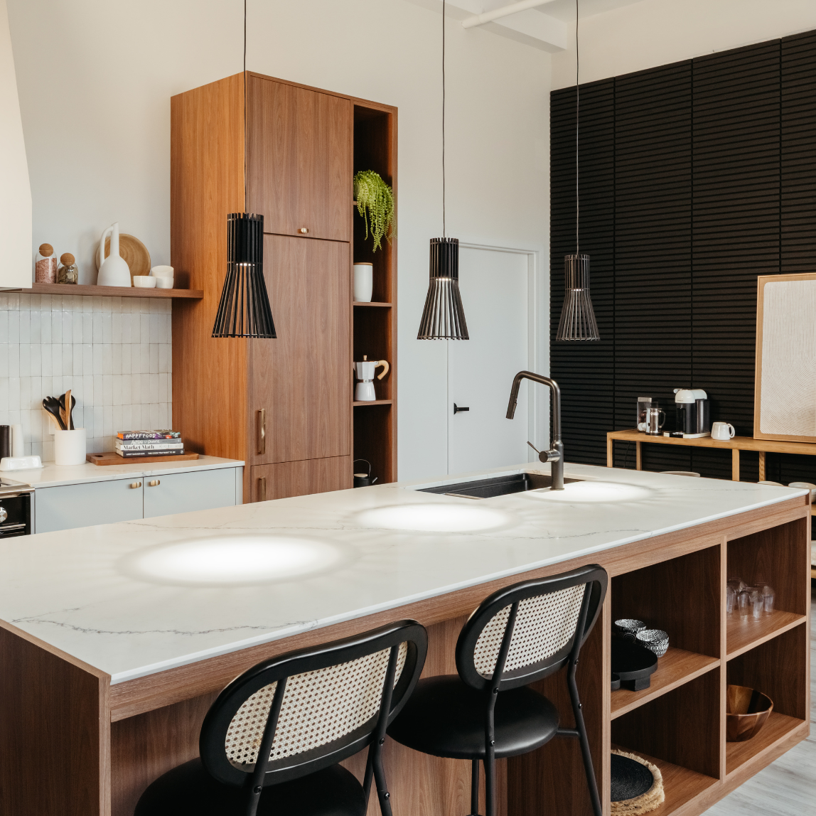 Modern kitchen with a marble island, two black chairs, wooden cabinets, black pendant lights, a built-in sink, and shelves with kitchen items. Black wall panels and decor accents complete the contemporary look.
