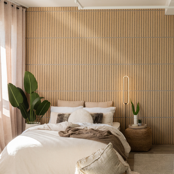 A cozy bedroom with a bed covered in white bedding and beige pillows, a woven ottoman nightstand, two potted green plants, and a modern light fixture against a wooden slat accent wall. Soft sunlight filters through curtains.