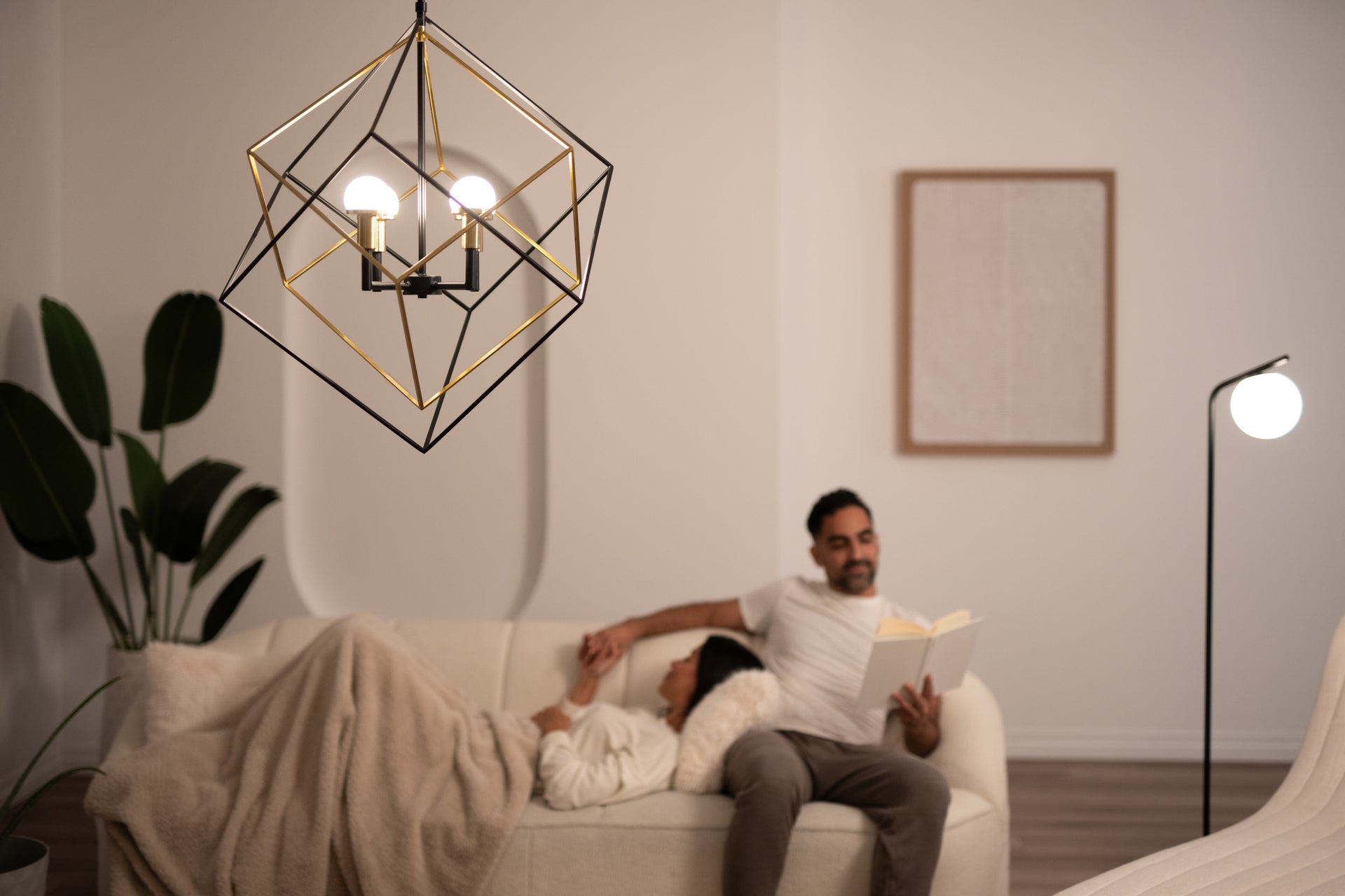 A modern geometric chandelier hangs in a cozy, softly lit living room. A couple relaxes on a white sofa; one person reads a book while the other rests with a blanket. A plant and a floor lamp decorate the space.